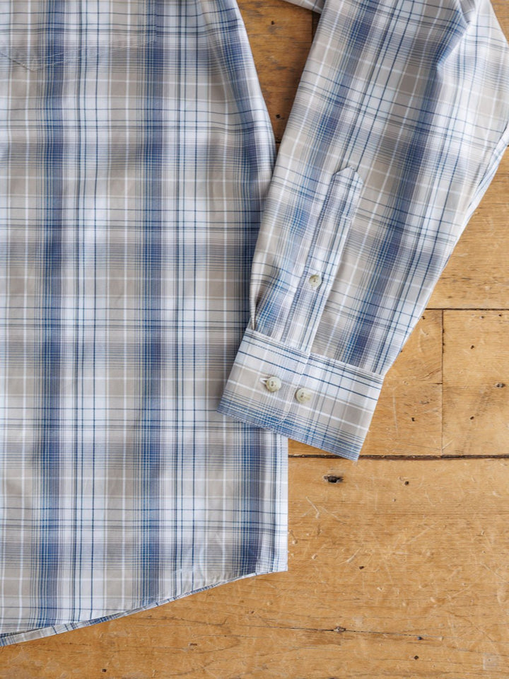 A close-up of the Wesson Ariat Men's Plaid Button Up by Savannah Sevens western life{&}style in blue, white, and beige, is laid flat on wood, showing buttoned cuffs and body. Features moisture wicking for comfort.