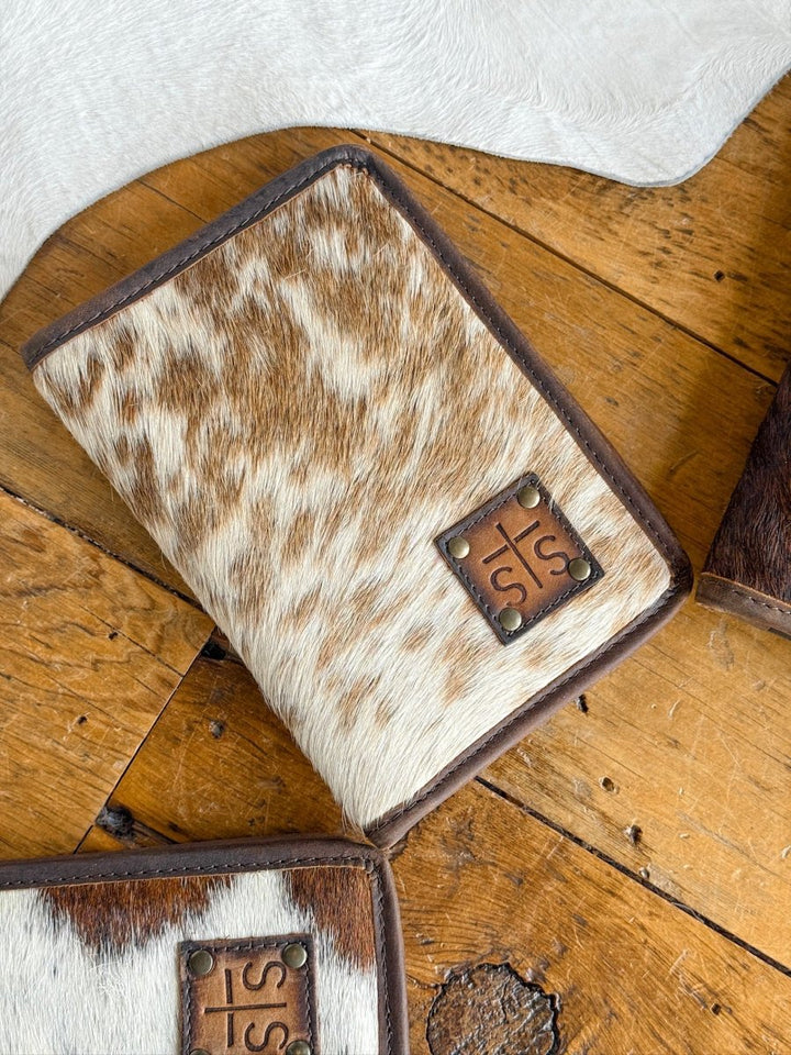 The West Tex Wallet by Savannah Sevens western life{&}style, featuring brown and white cowhide with leather accents and metal rivets, rests on a rustic wooden surface, partially overlapping another similar wallet.