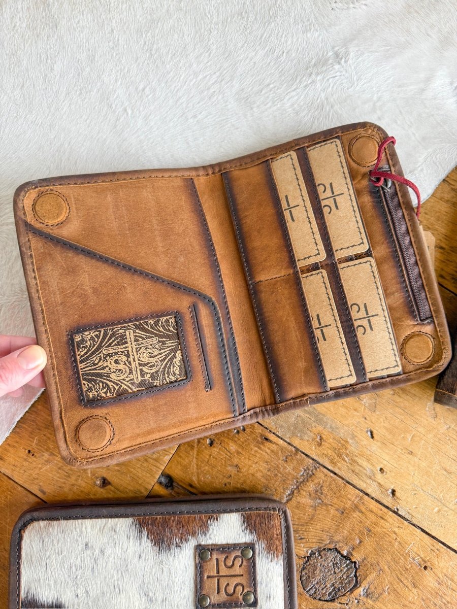 A hand holds open a Savannah Sevens West Tex Wallet in brown leather with card slots and cross-stamped cards, resting on wood beside an STS Ranch cowhide wallet featuring a leather patch.
