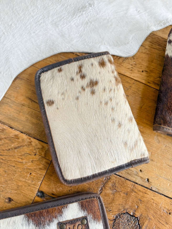 The West Tex Wallet by Savannah Sevens western life{&}style, in light brown and white cowhide with dark brown edges, rests on a wooden surface with other matching cowhide accessories.