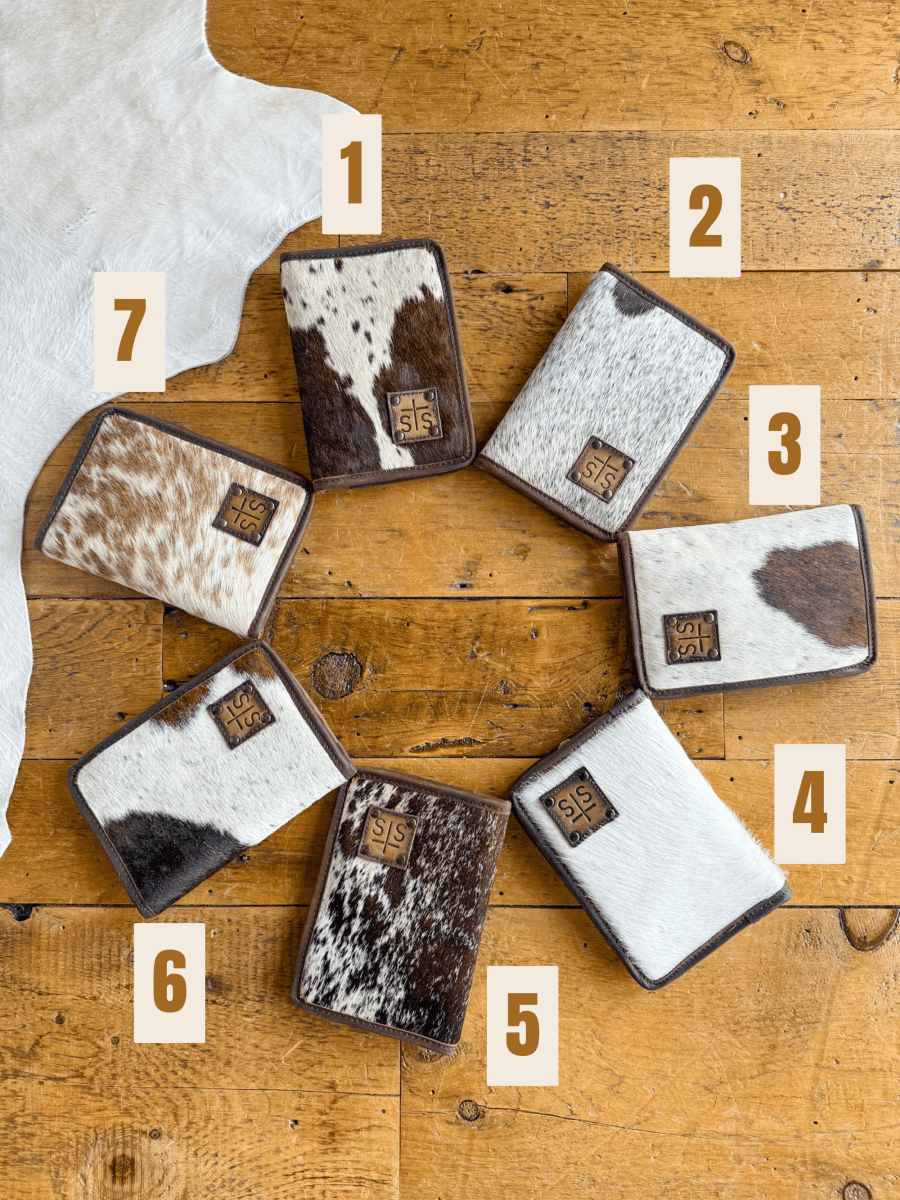 Seven West Tex Wallets by Savannah Sevens western life{&}style, each featuring a unique brown and white cowhide pattern and labeled 1 to 7, are arranged in a circle on wood. A partially visible wallet is in the top left corner.