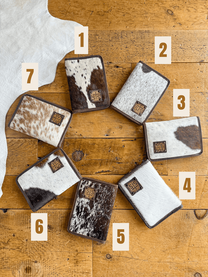 Seven West Tex Wallets by Savannah Sevens western life{&}style, each featuring a unique brown and white cowhide pattern and labeled 1 to 7, are arranged in a circle on wood. A partially visible wallet is in the top left corner.
