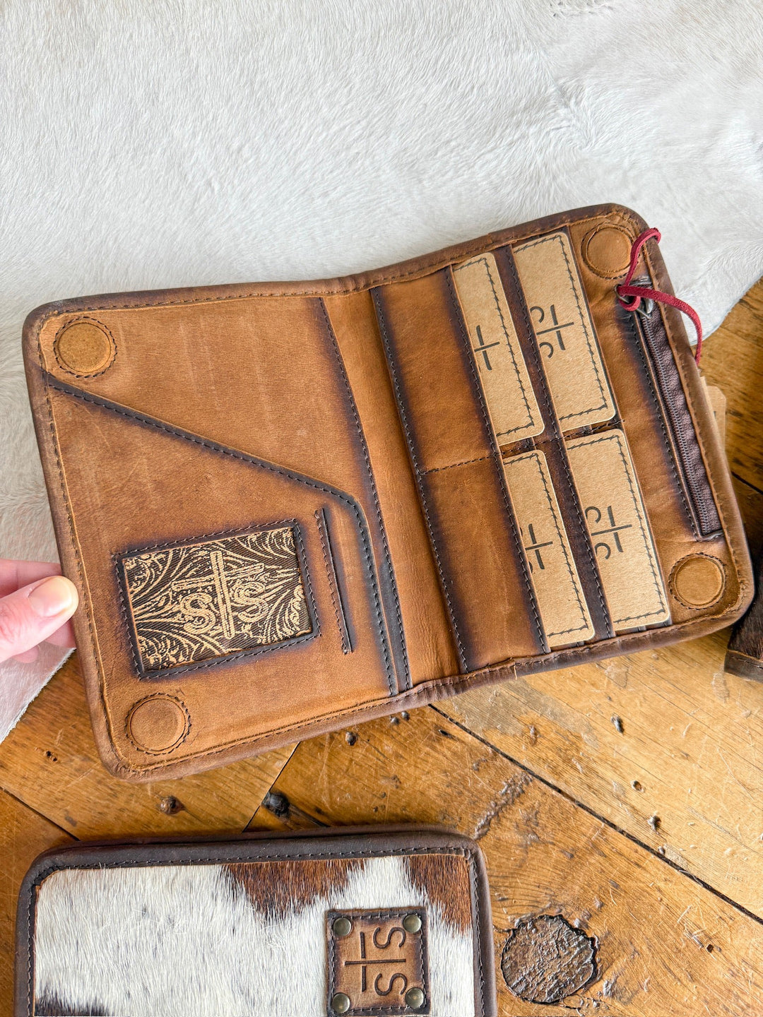 A hand holds open a Savannah Sevens West Tex Wallet in brown leather with card slots and cross-stamped cards, resting on wood beside an STS Ranch cowhide wallet featuring a leather patch.