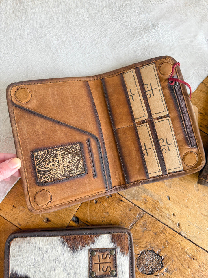 A hand holds open a Savannah Sevens West Tex Wallet in brown leather with card slots and cross-stamped cards, resting on wood beside an STS Ranch cowhide wallet featuring a leather patch.