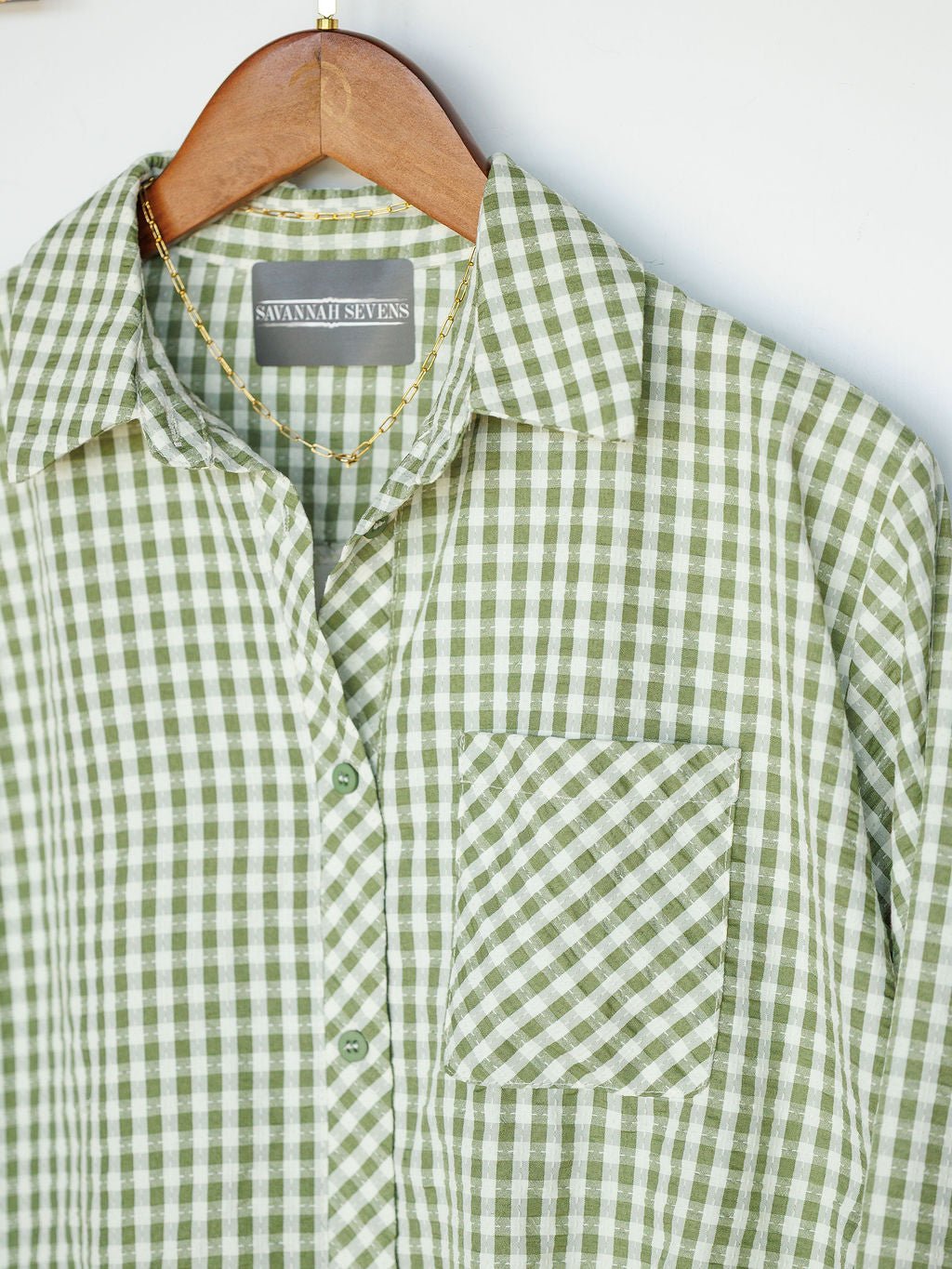 A green and white gingham button-up top called The Westbrook by Savannah Sevens western life{&}style hangs on a wooden hanger with a gold chain necklace around the collar.