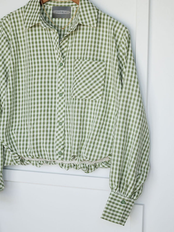 The Westbrook by Savannah Sevens western life{&}style is a green and white gingham button-up with a front pocket, relaxed fit, gathered hem, and subtle balloon sleeves, shown hanging on a white background.