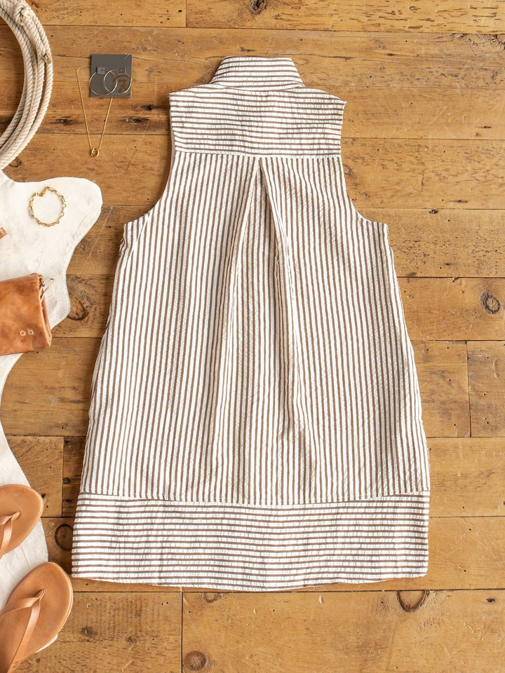 The Westerly Brown Striped Button Up Dress by Savannah Sevens western life{&}style—sleeveless and knee-length—lies flat on a wooden floor, surrounded by sandals, a bracelet, a small purse, and a pair of earrings.