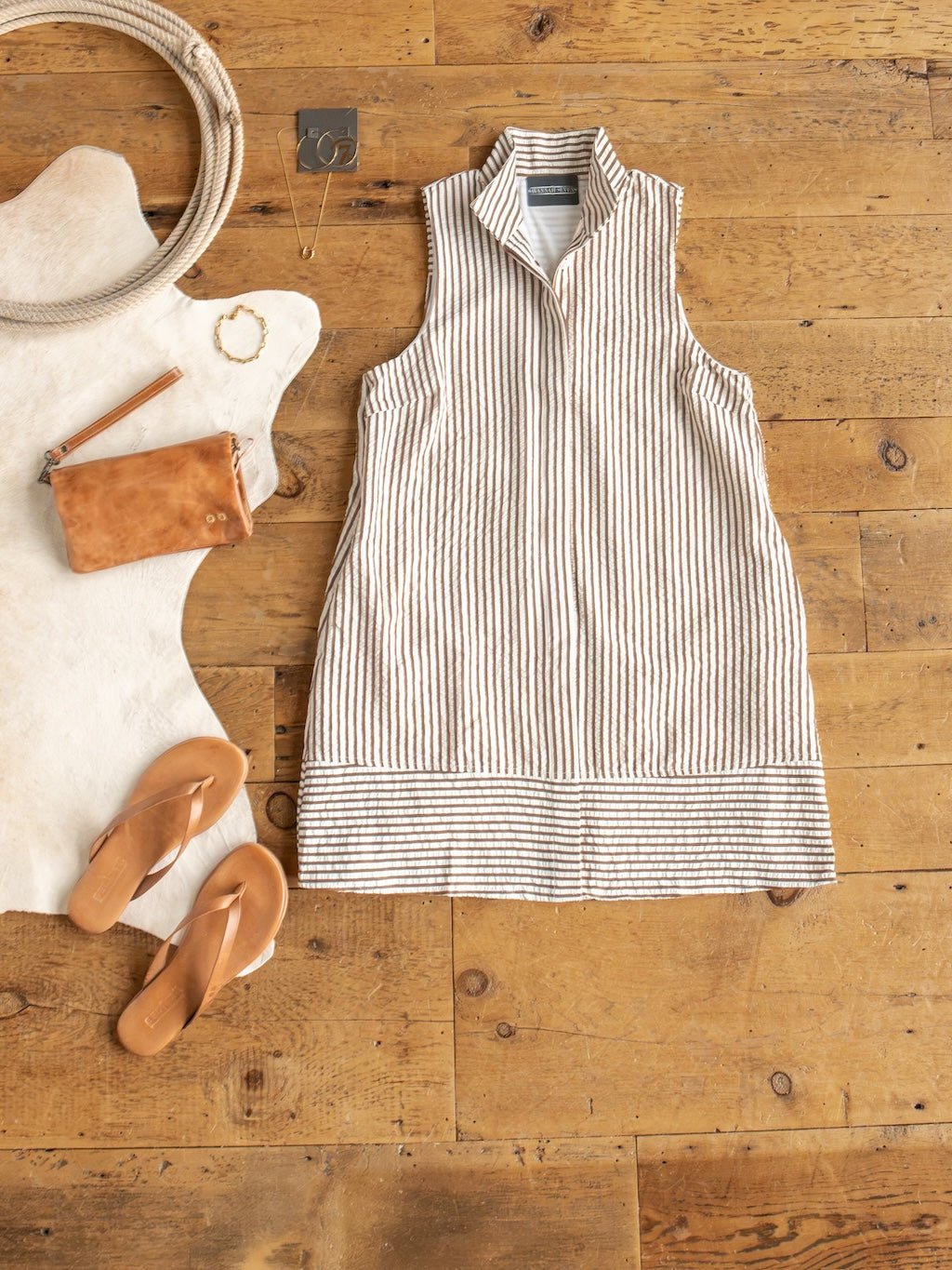 The Westerly Brown Striped Button Up Dress by Savannah Sevens western life{&}style is styled flat on a wooden floor, paired with tan sandals, a brown wristlet, jewelry accents, a coiled rope detail, and complemented by a white rug—showcasing an effortlessly casual summer outfit.
