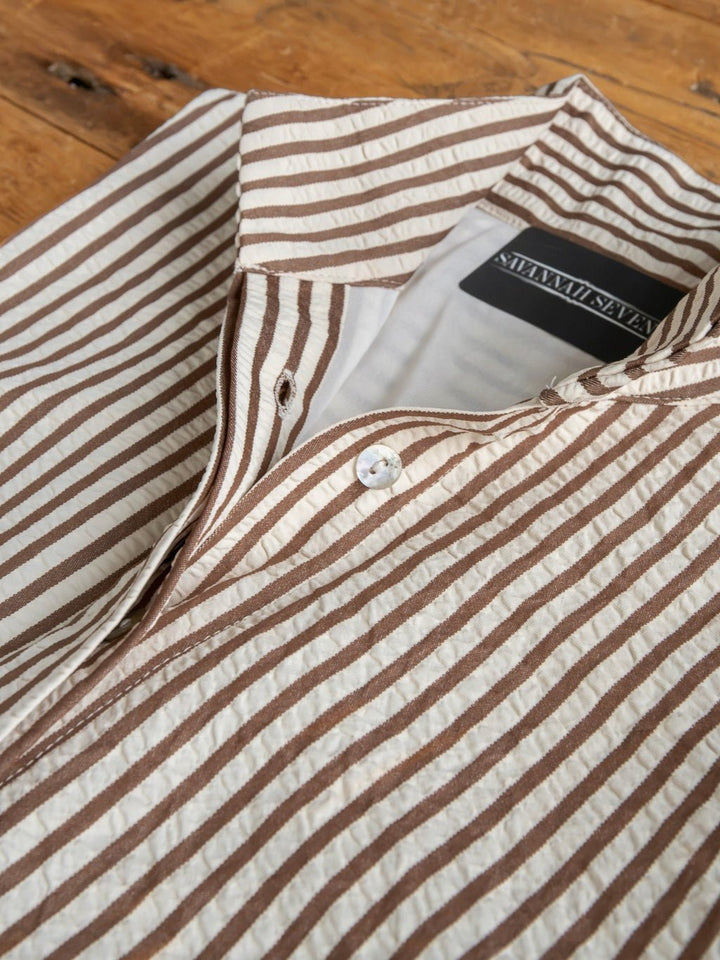 A close-up of the Westerly Brown Striped Button Up Dress by Savannah Sevens western life{&}style, featuring brown and cream vertical stripes, a visible white button, and a black brand label, laid flat on a wooden surface.
