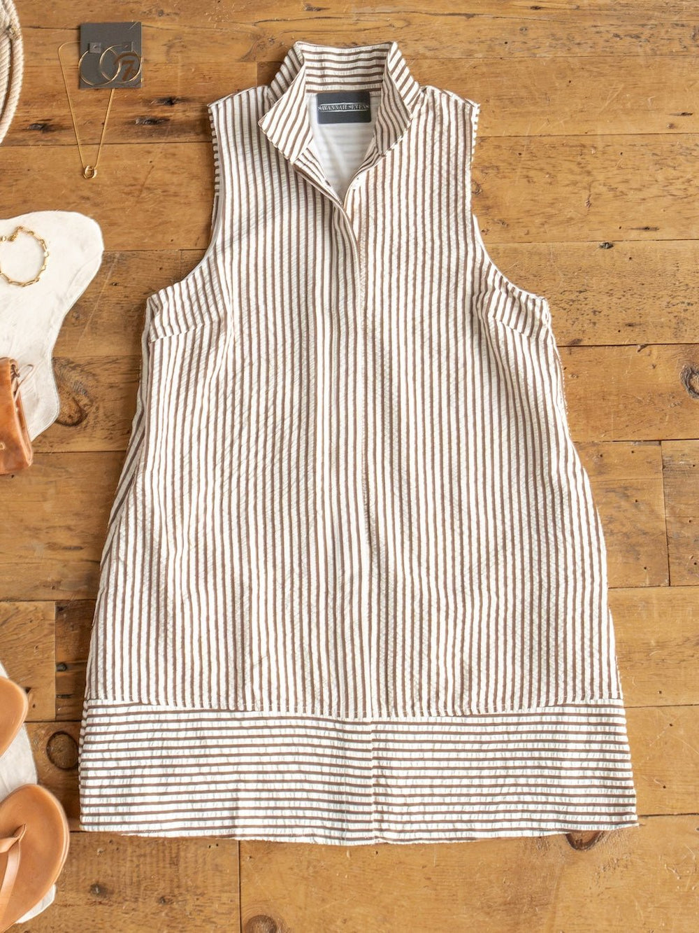 The Westerly Brown Striped Button Up Dress by Savannah Sevens western life{&}style, a linen sleeveless dress with vertical beige and white stripes, a high collar, and horizontal stripes at the hem, is laid flat on a wooden floor with jewelry and sandals arranged nearby.