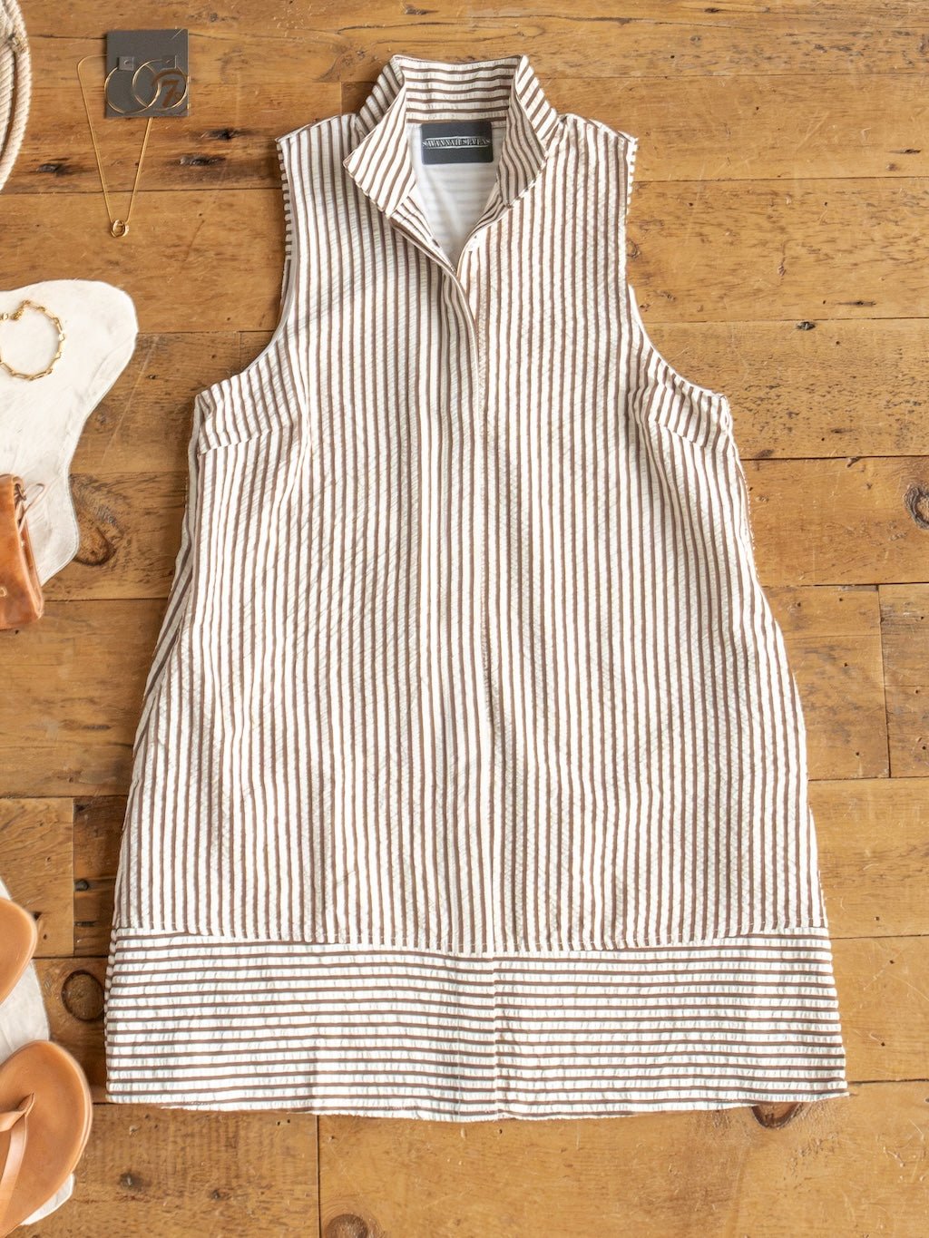 The Westerly Brown Striped Button Up Dress by Savannah Sevens western life{&}style, a linen sleeveless dress with vertical beige and white stripes, a high collar, and horizontal stripes at the hem, is laid flat on a wooden floor with jewelry and sandals arranged nearby.