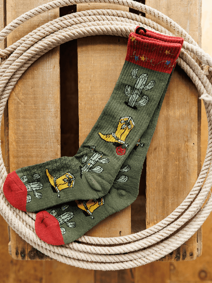 The Savannah Sevens western life{&}style Western Lights Crew Socks, in green with red toes and cuffs and cactus and cowboy boot designs, are made from an odor-fighting hemp blend for lightweight breathability. Displayed inside a coiled rope on wood.