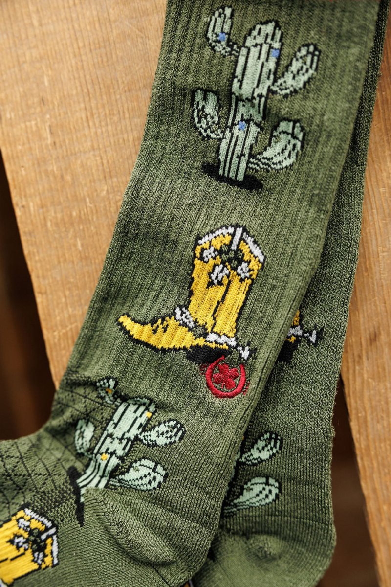 Western Lights Crew Socks by Savannah Sevens western life{&}style, featuring yellow cowboy boots, green cacti, and red spurs on a hemp blend for odor control, are shown draped over a wooden surface.