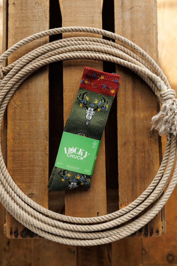 A pair of Western Lights Crew Socks by Savannah Sevens western life{&}style, featuring a green cactus and flower pattern, rests inside a coiled rope on wood. These lightweight, breathable socks are designed for Western performance.