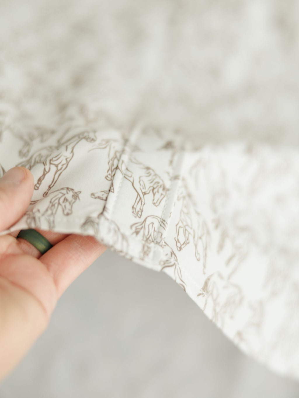 A hand holds the edge of white fabric with a light brown horse print from the Weston Wrangler Horse Button Top by Savannah Sevens western life{&}style, ideal for a women's western shirt; the softly focused image highlights the pattern.