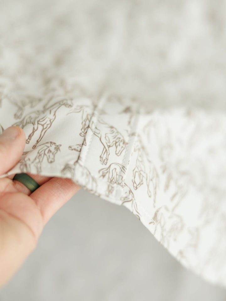 A hand holds the edge of white fabric with a light brown horse print from the Weston Wrangler Horse Button Top by Savannah Sevens western life{&}style, ideal for a women's western shirt; the softly focused image highlights the pattern.