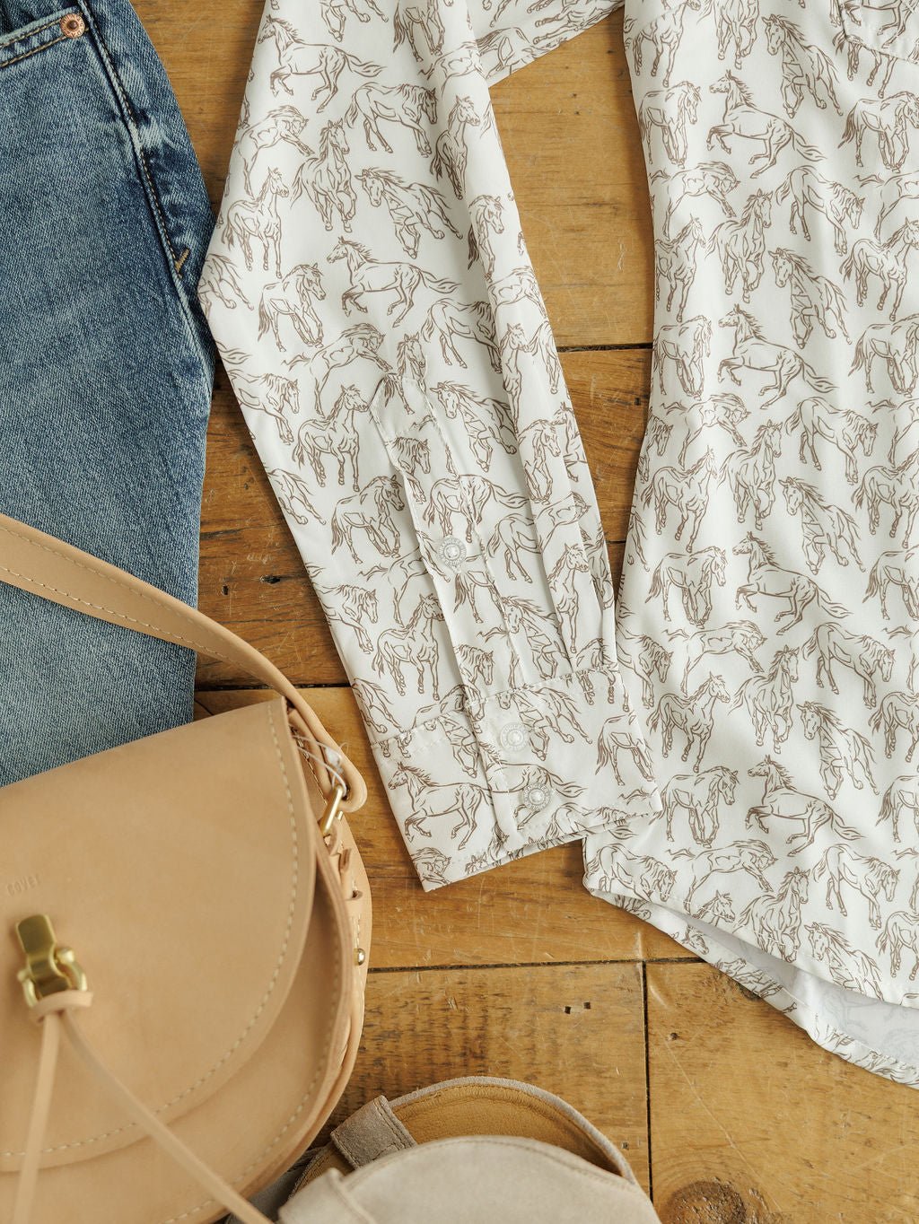 A flat lay featuring Savannah Sevens western life{&}style's Weston Wrangler Horse Button Top, blue jeans, a beige leather crossbody bag, and light-colored shoes on a wooden floor.
