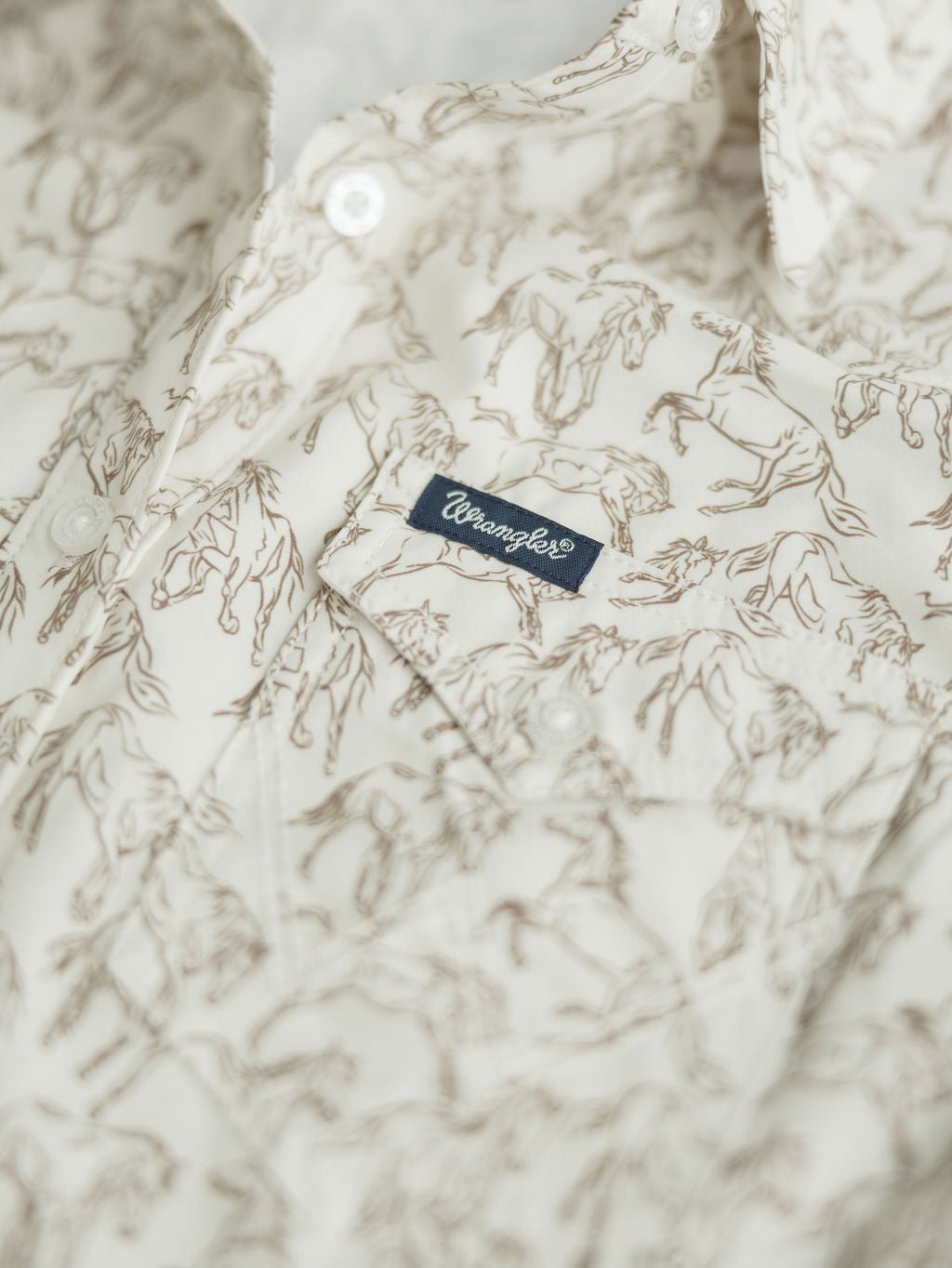 Close-up of the Weston Wrangler Horse Button Top by Savannah Sevens western life{&}style, featuring a brown horse print, small blue Wrangler® tag on the chest pocket, and detailed white buttons.