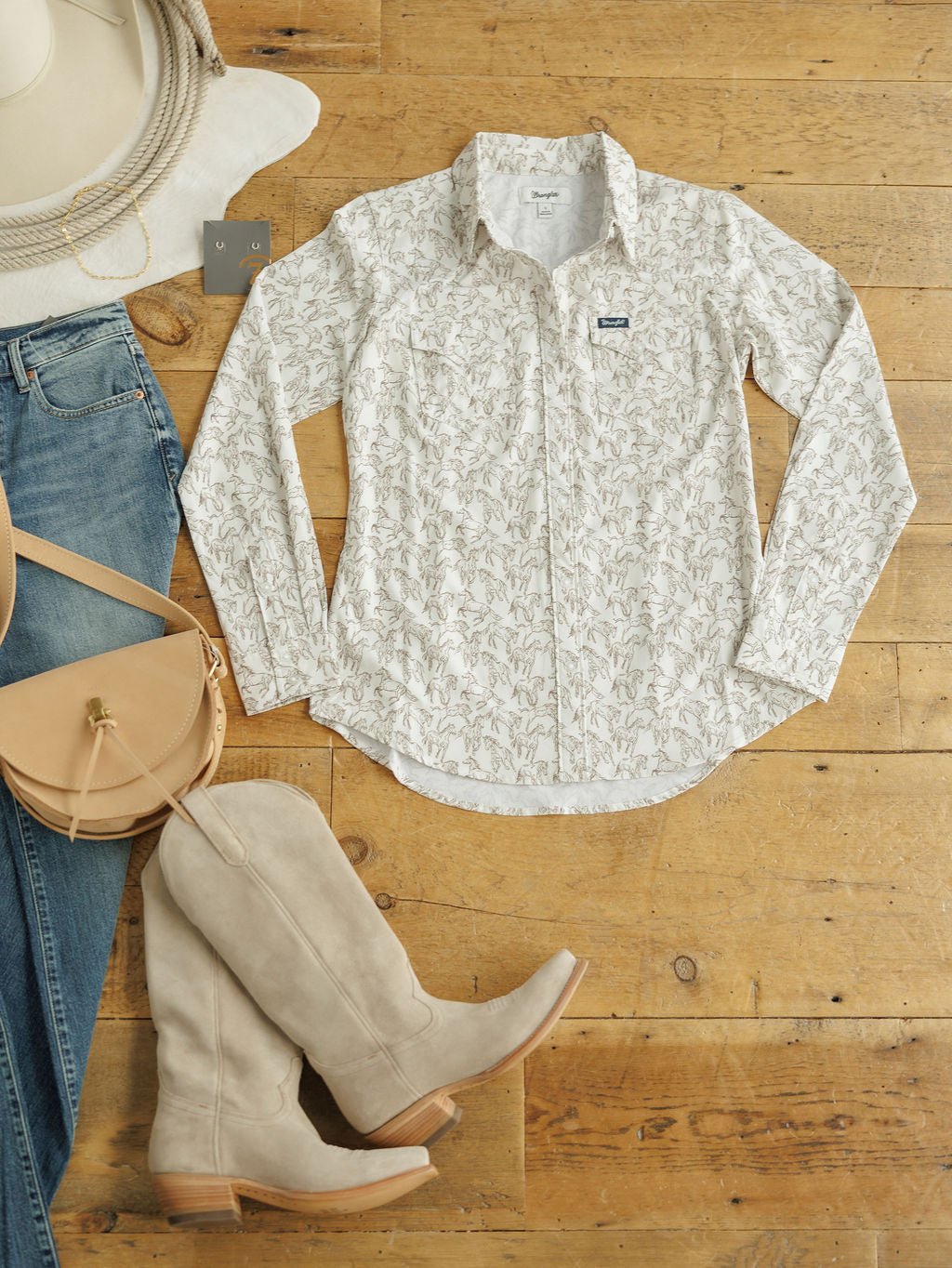 The Weston Wrangler Horse Button Top by Savannah Sevens western life{&}style is styled on a wooden floor with light blue jeans, beige knee-high boots, a tan crossbody bag, a white hat, and silver earrings.