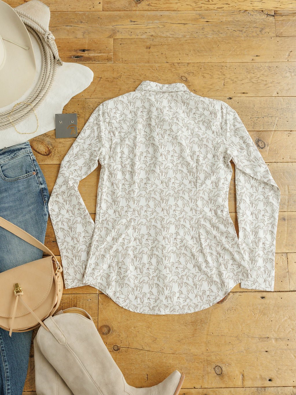 The Weston Wrangler Horse Button Top by Savannah Sevens western life{&}style, a long-sleeve white women’s shirt with a subtle beige floral pattern, is displayed flat with a beige hat, blue jeans, tan purse and boots, and a belt.