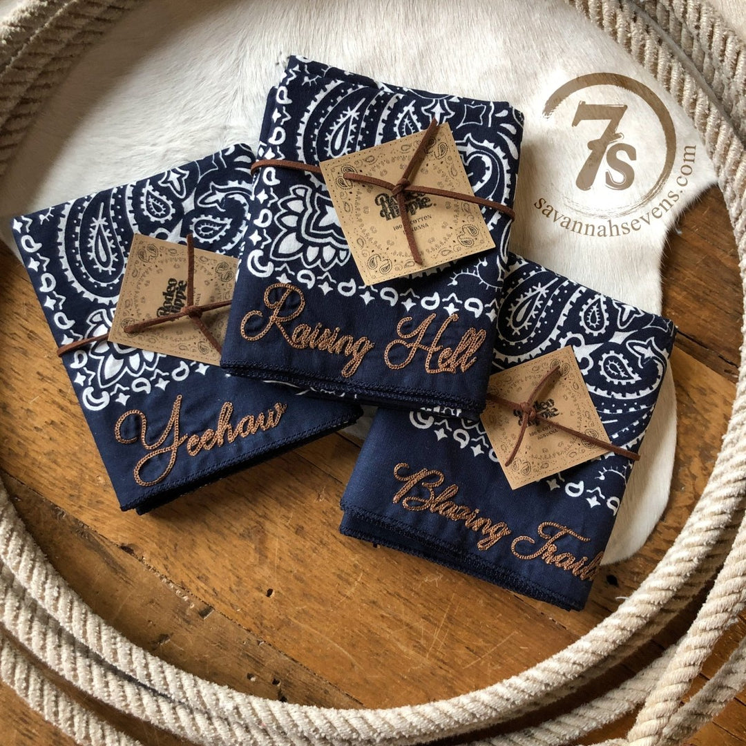 The Westover Bandana Scarf by Savannah Sevens western life{&}style features three cotton navy color wash bandanas with white paisley, each embroidered with Yeehaw, Raising Hell, or Chasing Trouble and tied with leather cords. Logo included nearby.