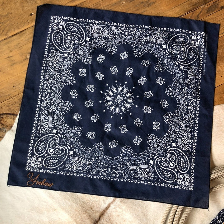 The Westover Bandana Scarf by Savannah Sevens western life{&}style, featuring a navy color wash with white paisley and floral patterns and "Yeehaw" embroidered in orange script, is laid flat on wood with a rope and tan rug partially visible underneath.