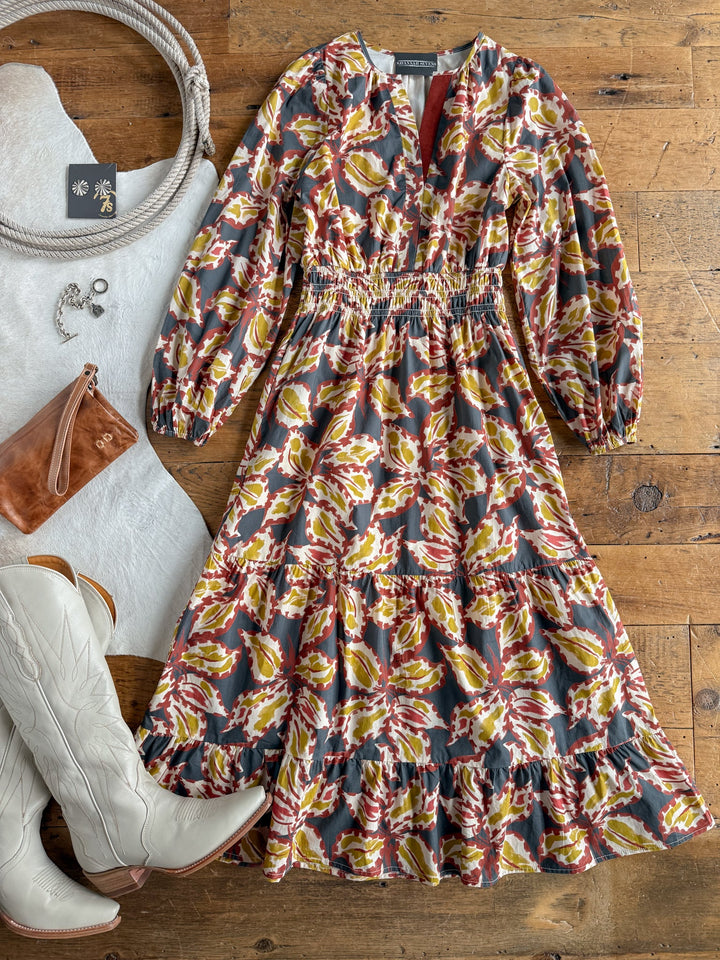 The Westphalia Floral Dress by Savannah Sevens western life{&}style is styled with white cowboy boots, a brown clutch, jewelry, a coiled rope, and a white fringed fabric—all displayed on a rustic wooden surface.