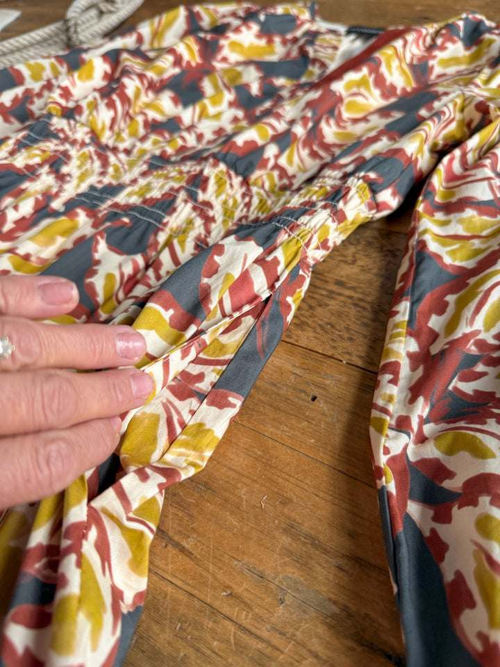 A hand with a ring touches the Westphalia Floral Dress by Savannah Sevens western life{&}style, featuring a red, yellow, and white floral print on a gray background, as it lies on a wooden surface.