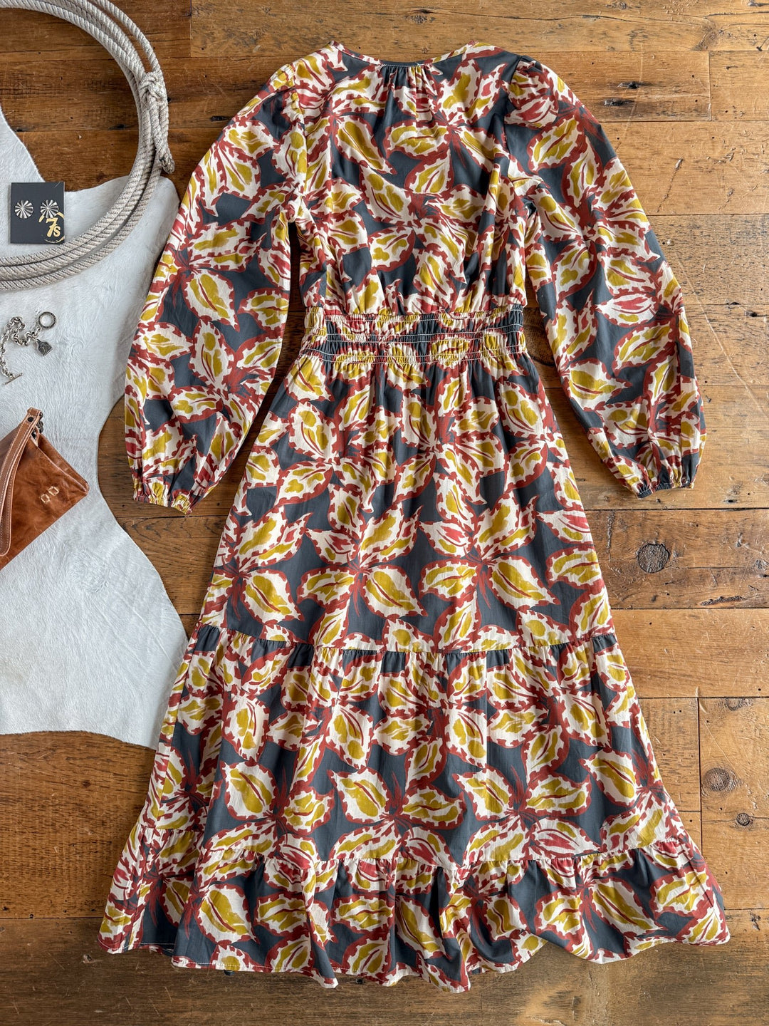 The Westphalia Floral Dress by Savannah Sevens western life{&}style, featuring earthy-toned florals in a midi length, is displayed on a wooden floor. Nearby are a tan purse, a white cloth, a coiled rope, and a small black card.