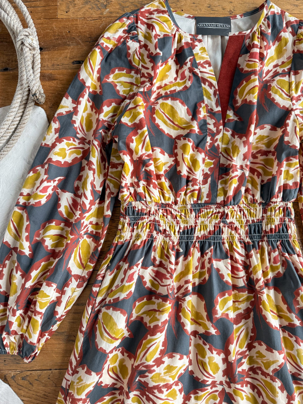 The Westphalia Floral Dress by Savannah Sevens western life{&}style, featuring a red, yellow, and white floral pattern on a gray cotton fabric with long sleeves, is displayed on a wooden surface next to a coiled rope.