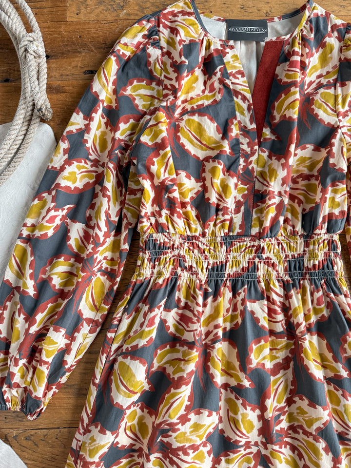 The Westphalia Floral Dress by Savannah Sevens western life{&}style, featuring a red, yellow, and white floral pattern on a gray cotton fabric with long sleeves, is displayed on a wooden surface next to a coiled rope.
