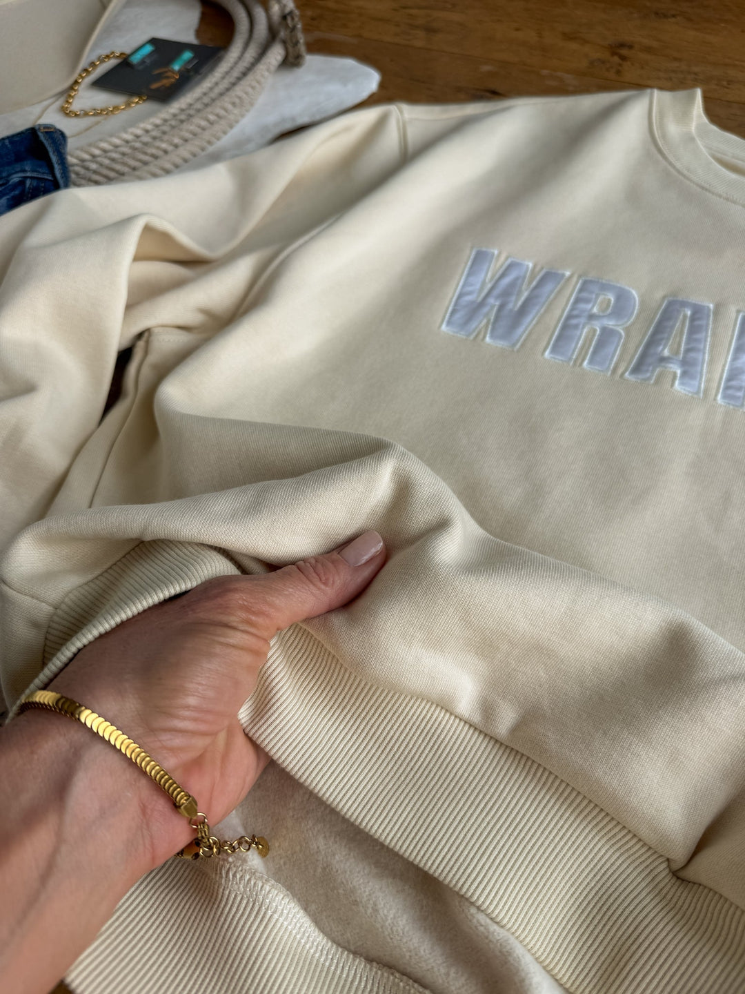 A hand adorned with a gold bracelet holds the Westport Cream Pullover by Savannah Sevens western life{&}style, a cream-colored cotton blend sweatshirt featuring raised white “WRAN” lettering, displayed on a wooden surface with other relaxed fit apparel visible nearby.
