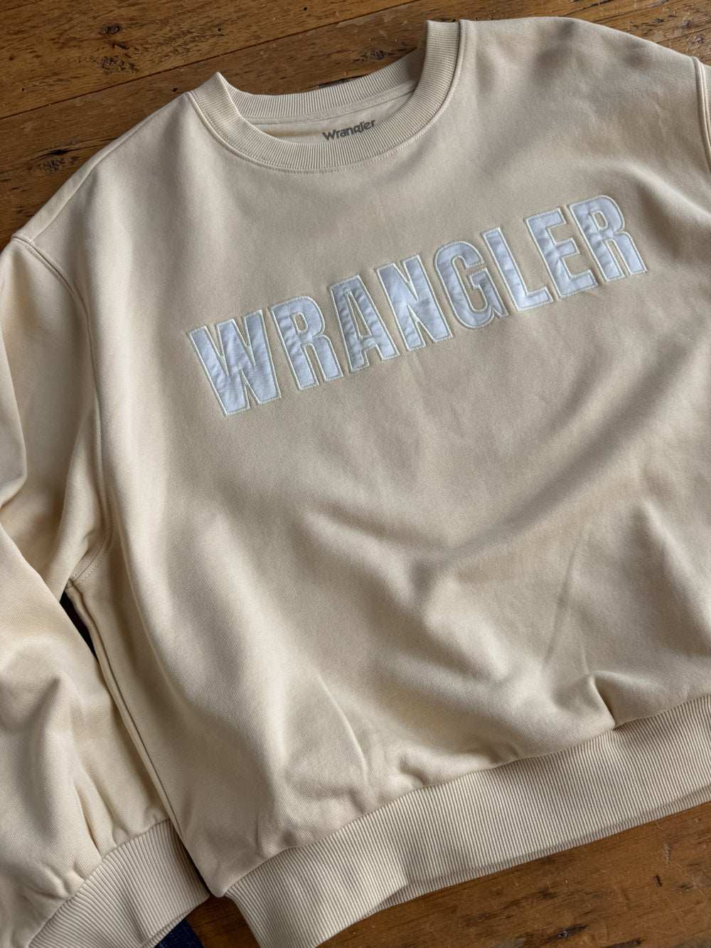 The Westport Cream Pullover by Savannah Sevens western life{&}style is a beige graphic logo sweatshirt featuring “WRANGLER” in large white letters across the chest. Crafted from a soft cotton blend for a relaxed fit, it is shown laid flat on a wooden surface.