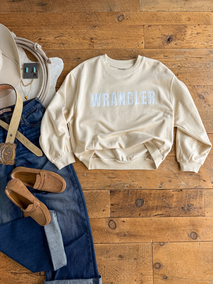 A Westport Cream Pullover by Savannah Sevens western life{&}style in a relaxed fit is laid on a wooden floor, surrounded by blue jeans, a tan belt, brown loafers, a white cowboy hat, a rope, and some jewelry.