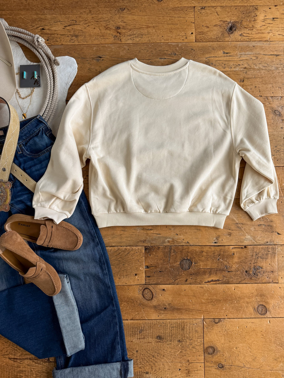 The Westport Cream Pullover by Savannah Sevens western life&style, a cotton-blend crewneck sweatshirt, is laid flat on a wooden floor alongside blue jeans, tan loafers, a belt, a necklace, and a white hat.