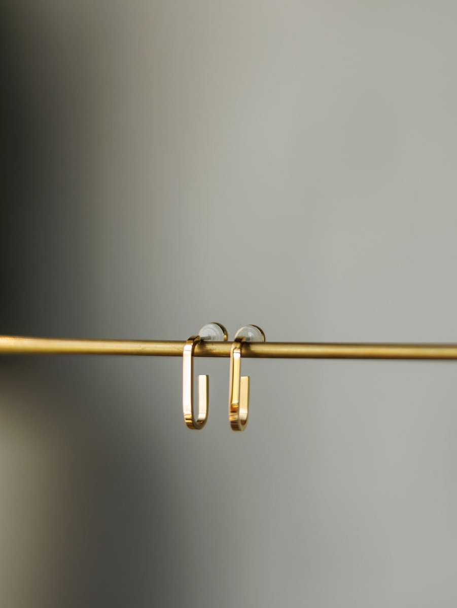 The Westport Earrings by Savannah Sevens western life{&}style, minimalist 18K gold plated hoops, hang on a thin gold rod against a blurred gray background.