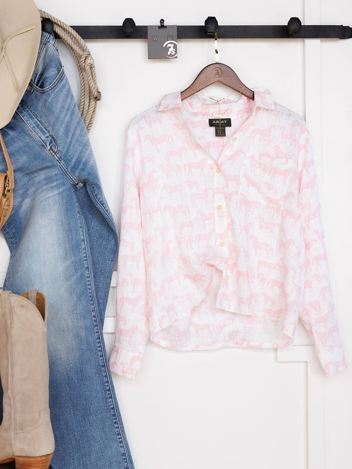 The Westview Horse Graphic Button Up Top by Savannah Sevens western life{&}style, in light pink and white with a horse print, hangs beside blue jeans and a beige bag on hooks against a white wall.
