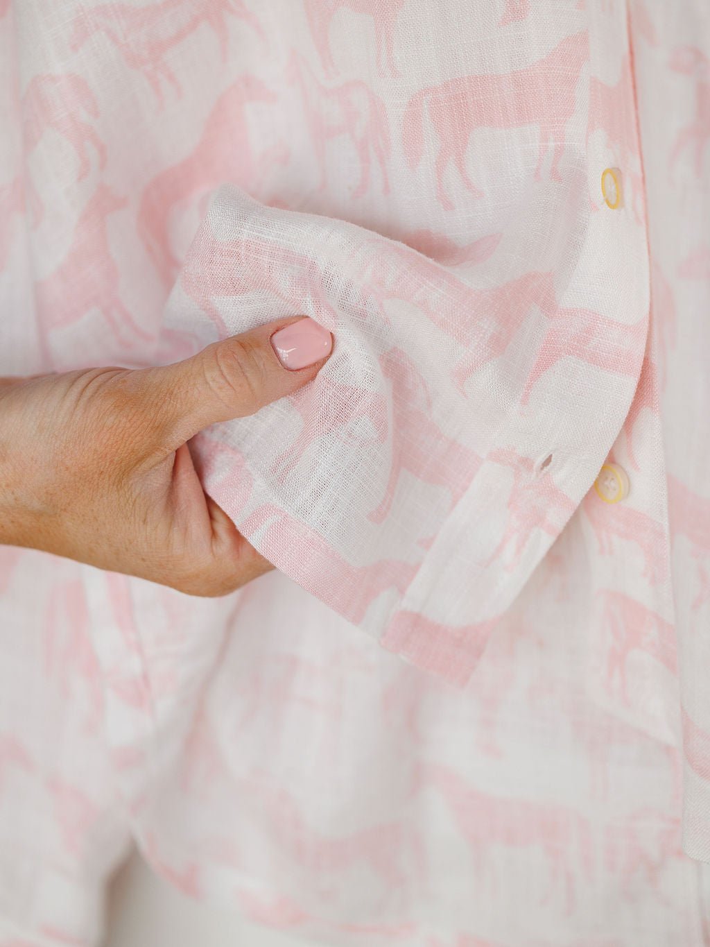 A hand with light pink nail polish holds the Westview Horse Graphic Button Up Top by Savannah Sevens western life{&}style, featuring a white and pink horse print and yellow buttons.