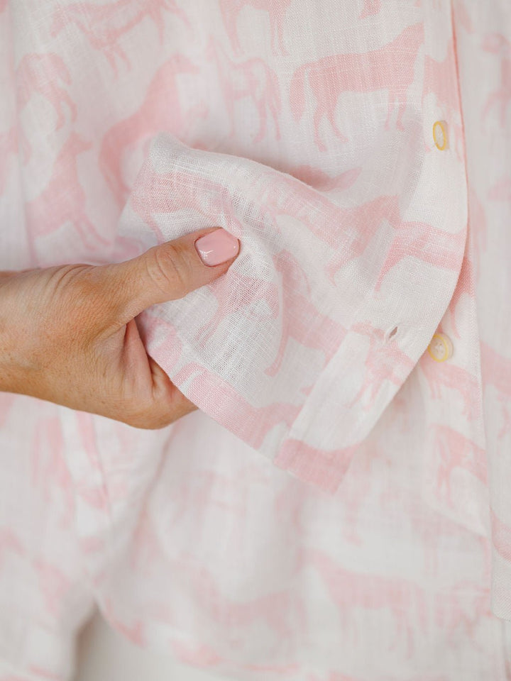 A hand with light pink nail polish holds the Westview Horse Graphic Button Up Top by Savannah Sevens western life{&}style, featuring a white and pink horse print and yellow buttons.