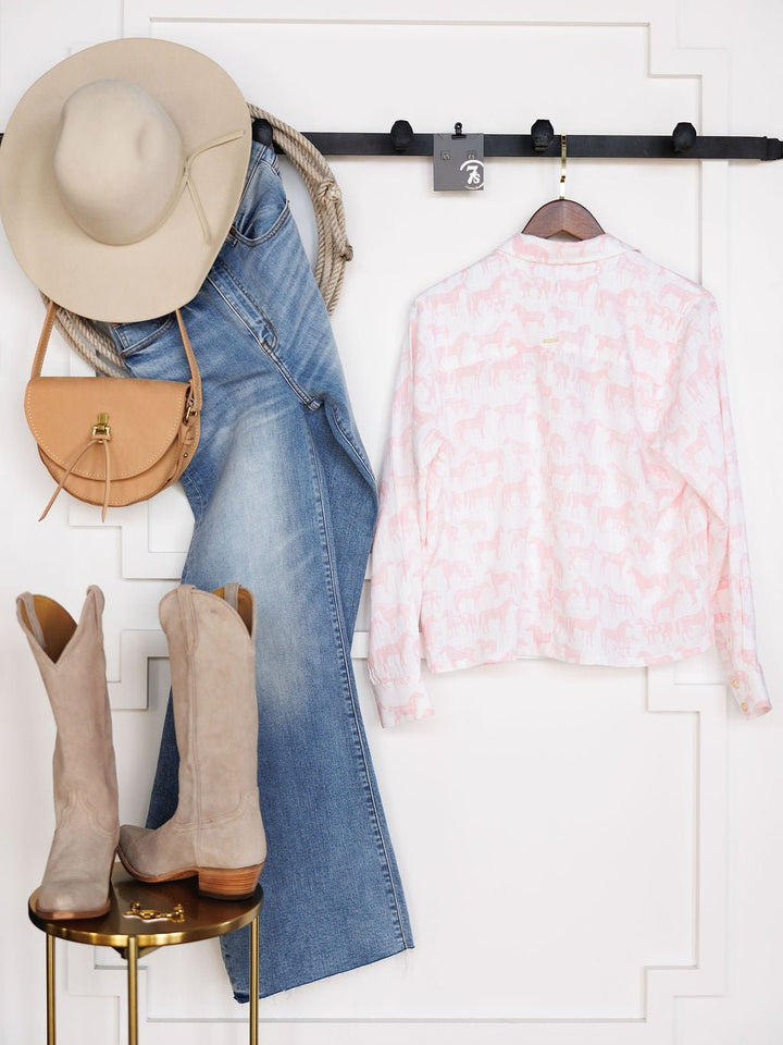 A Western-inspired look features a beige wide-brim hat, light brown saddle bag, blue flared jeans, Savannah Sevens western life{&}style Westview Horse Graphic Button Up Top, and taupe knee-high boots against a white wall.