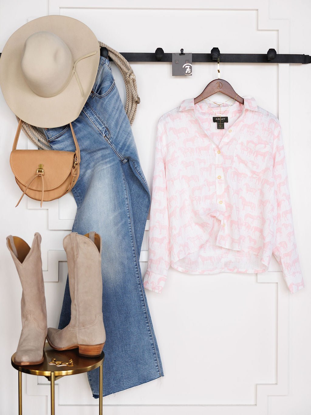 A beige hat, brown crossbody bag, and light blue jeans hang next to a Savannah Sevens western life{&}style Westview Horse Graphic Button Up Top in pink and white, while beige cowboy boots rest on a stool underneath.