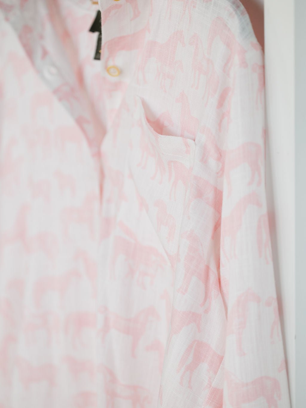 A close-up of the Westview Horse Graphic Button Up Top by Savannah Sevens western life{&}style, showing its light pink and white horse print, long sleeves, pocket, and a collar button, hanging on a hanger.
