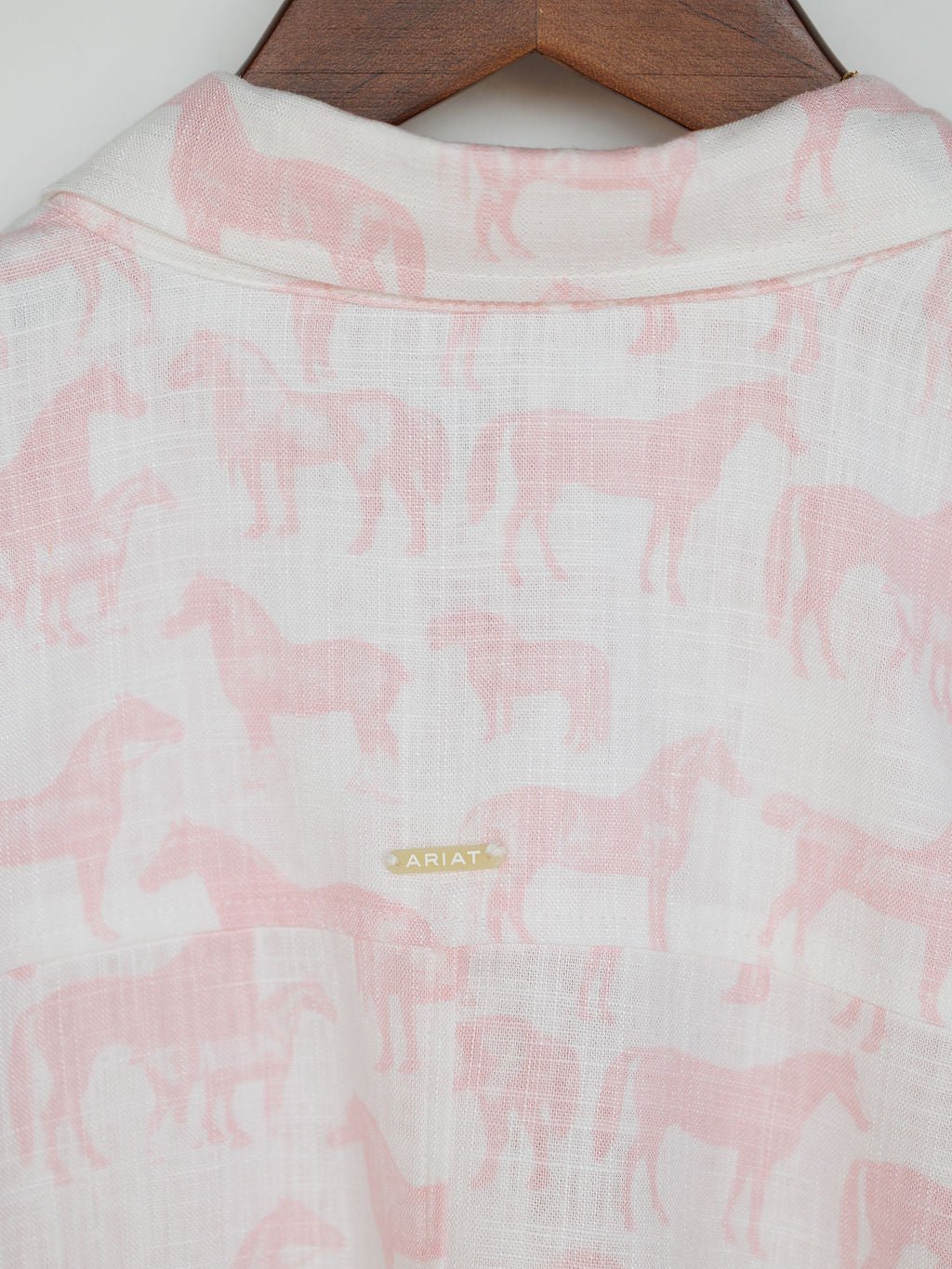 A close-up of the Westview Horse Graphic Button Up Top by Savannah Sevens western life{&}style, featuring a linen blend with pink horse print and a small gold label on the back, displayed on a wooden hanger.