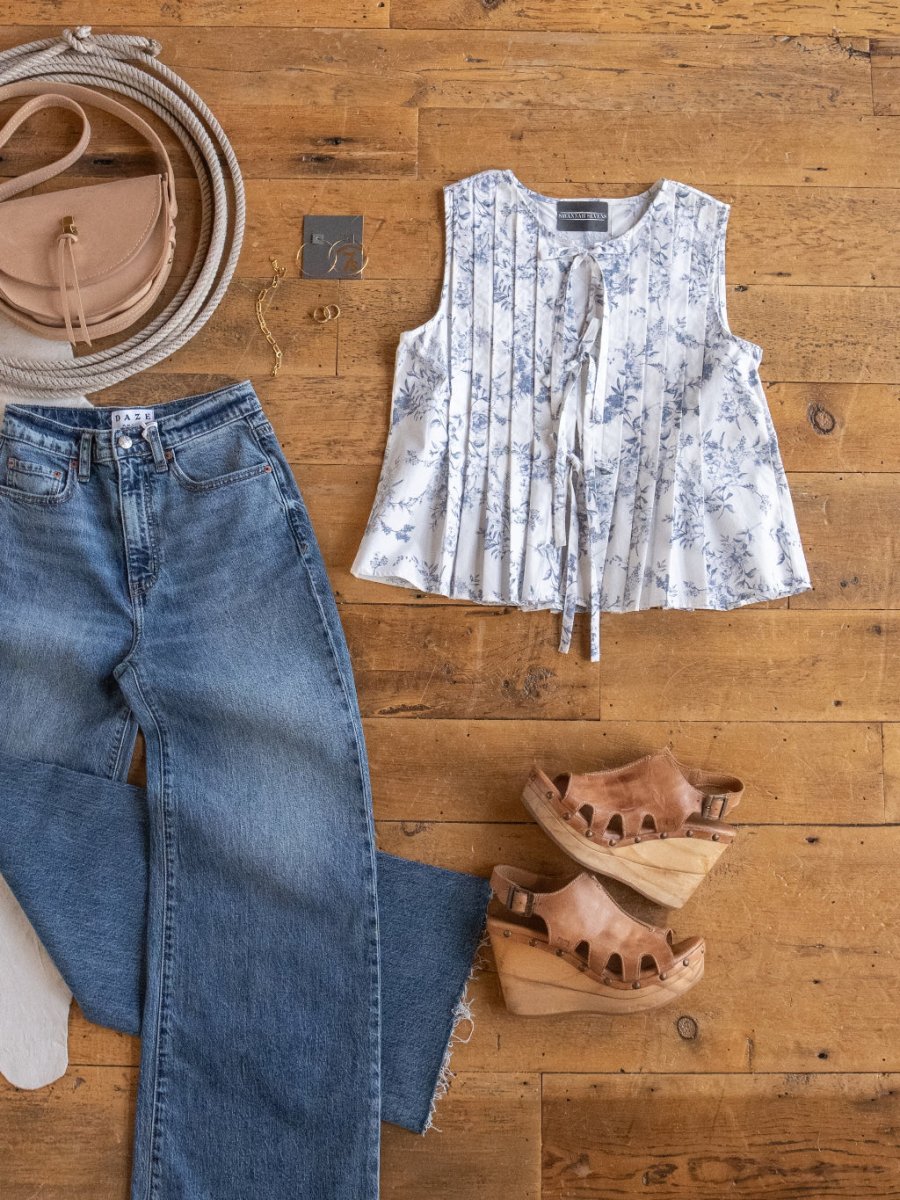 A flat lay of casual women’s clothing: wide-leg blue jeans, the Savannah Sevens western life{&}style Westwater Floral Tie Front Top, tan platform sandals, a round beige crossbody bag, and a coiled rope on a wooden floor.