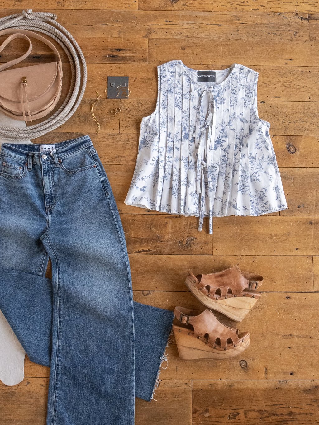 A flat lay of casual women’s clothing: wide-leg blue jeans, the Savannah Sevens western life{&}style Westwater Floral Tie Front Top, tan platform sandals, a round beige crossbody bag, and a coiled rope on a wooden floor.