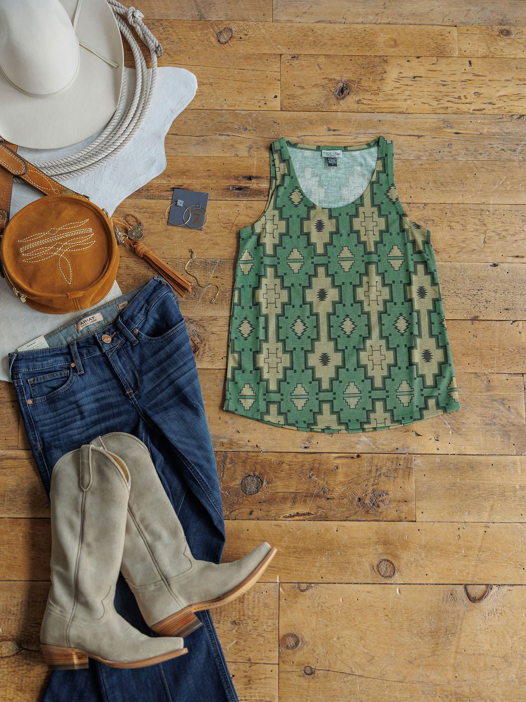 A flat lay on a wooden floor featuring the Savannah Sevens western life{&}style Wetonka Southwest Tank in green and beige, blue jeans, light gray cowboy boots, a tan round bag, and a white hat.