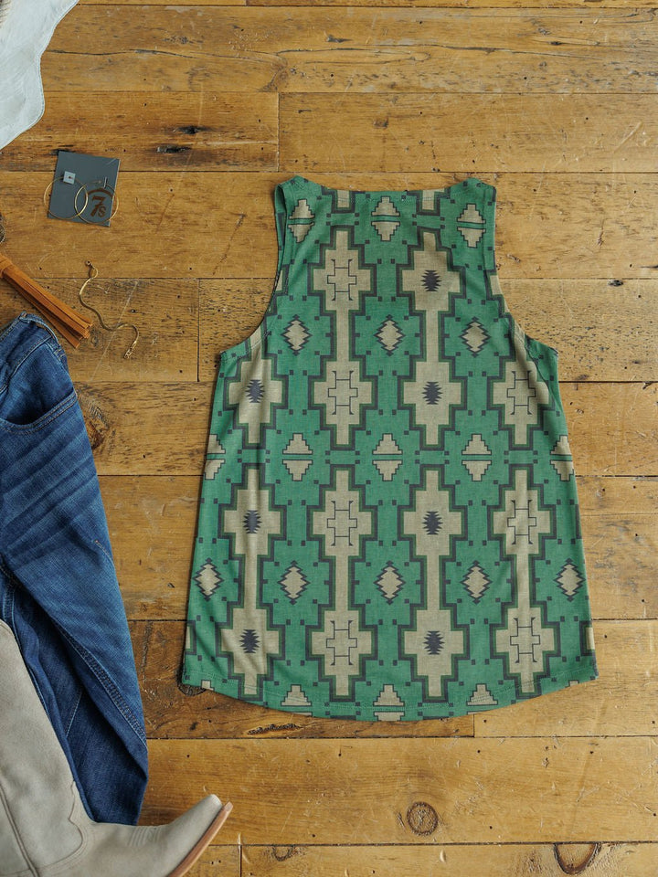 The Wetonka Southwest Tank by Savannah Sevens western life{&}style, featuring a green and beige geometric Southwest print, is displayed flat on a wooden floor beside blue jeans, taupe boots, and a young contemporary size tag.
