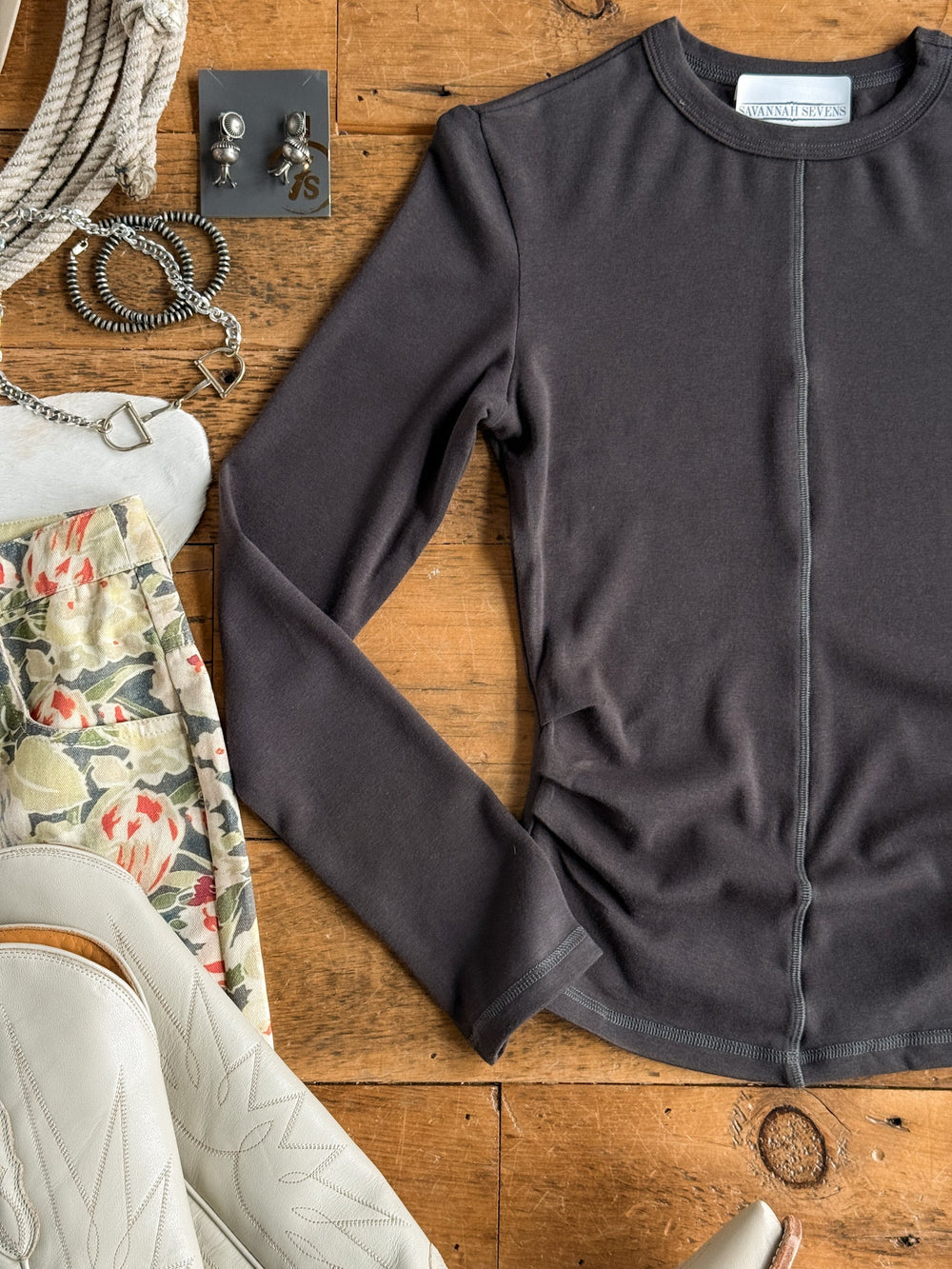 The Whitwell Charcoal Ruched Top by Savannah Sevens western life{&}style is displayed on a wooden surface, surrounded by earrings, bracelets, a rope, a floral skirt, and white cowboy boots.