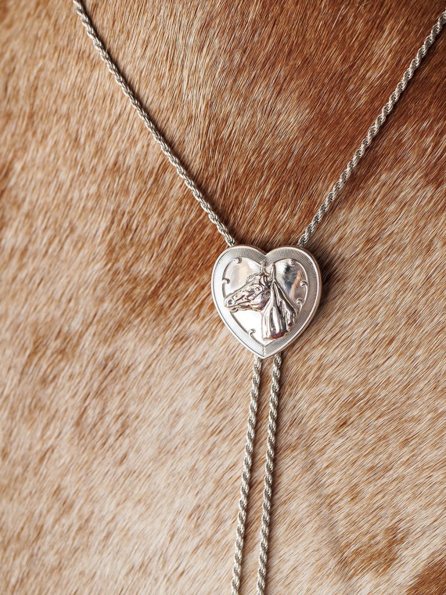 The Wild At Heart Bolo by Savannah Sevens western life{&}style features a sterling silver heart pendant with a raised horse design, shown resting on light brown, speckled cowhide—a bold Western jewelry statement.
