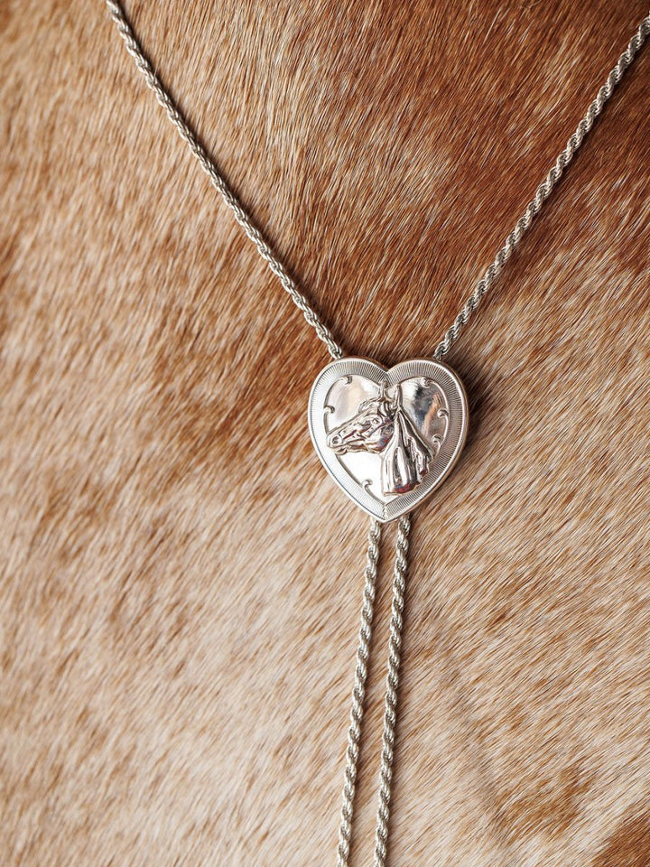 The Wild At Heart Bolo by Savannah Sevens western life{&}style features a sterling silver heart pendant with a raised horse design, shown resting on light brown, speckled cowhide—a bold Western jewelry statement.